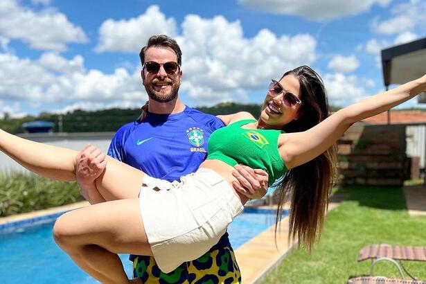 Laís Caldas sorrindo, com camisa regata verde, com uma bandeira do Brasil do lado esquerdo da camisa, short branco, cabelo solto, com braços abertos e um óculos de sol; Gustavo Marsengo sorrindo, de óculos de sol, camisa azul com brasão da CBF do lado esquerda da camisa e short estampado com bandeiras do Brasil em frente a uma piscina