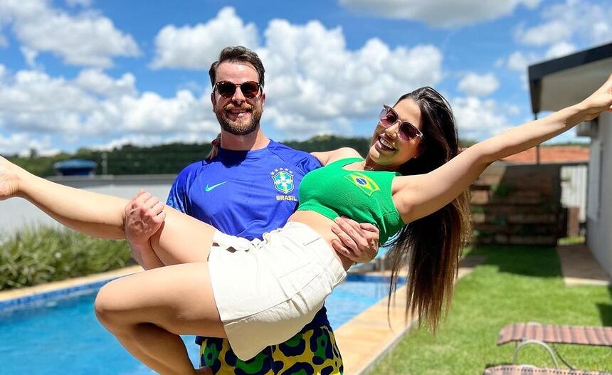 Laís Caldas sorrindo, com camisa regata verde, com uma bandeira do Brasil do lado esquerdo da camisa, short branco, cabelo solto, com braços abertos e um óculos de sol; Gustavo Marsengo sorrindo, de óculos de sol, camisa azul com brasão da CBF do lado esquerda da camisa e short estampado com bandeiras do Brasil em frente a uma piscina