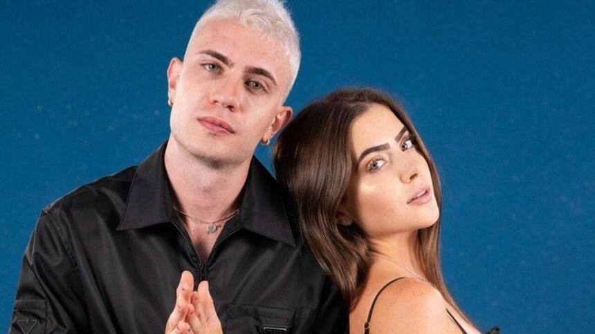 Leo Picon com camisa social preta, com as mãos juntas ao lado de Jade Picon, de perfil, cabelo castanho, de perfil, de vestido preto durante ensaio fotográfico publicitário