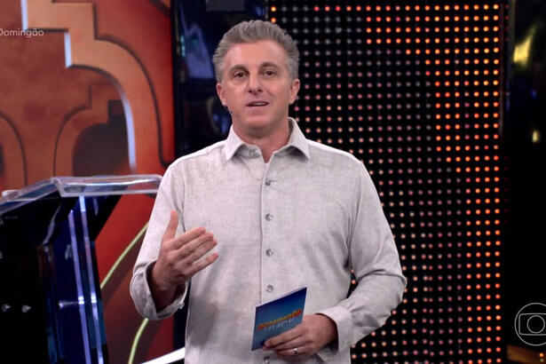 Luciano Huck, com uma blusa em tom claro, em trecho do Domingão, gesticulando com uma das mãos e segurando a ficha do programa com a outra