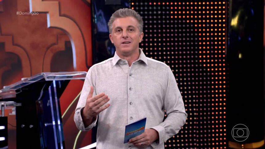 Luciano Huck, com uma blusa em tom claro, em trecho do Domingão, gesticulando com uma das mãos e segurando a ficha do programa com a outra