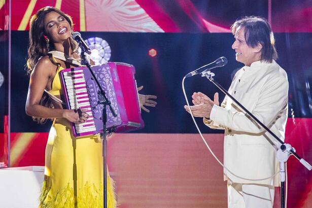 Lucy Alves de vestido amarelo, segurando uma sanfona ao lado de Roberto Carlos, que está de camisa e calça branca aplaudindo Lucy durante gravação de especial da TV Globo