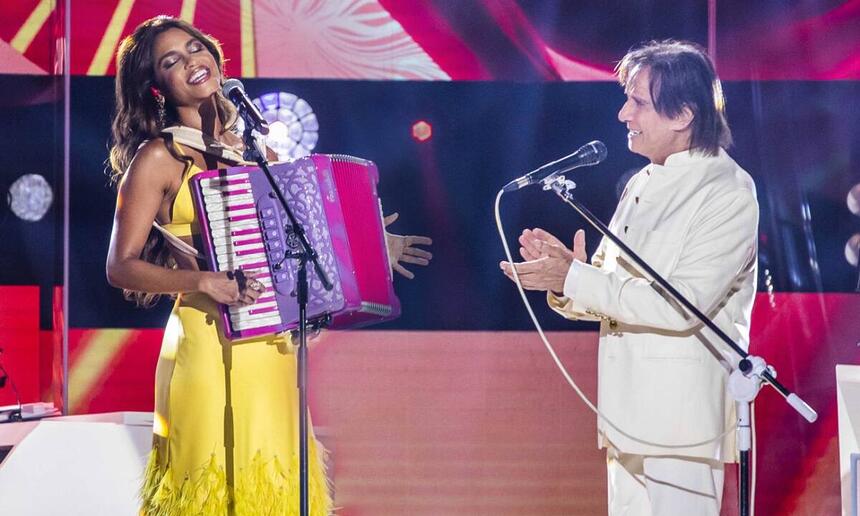 Lucy Alves de vestido amarelo, segurando uma sanfona ao lado de Roberto Carlos, que está de camisa e calça branca aplaudindo Lucy durante gravação de especial da TV Globo