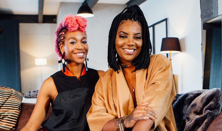 Ludmilla de roupão marrom, sorrindo, com cabelo trançado, ao lado de Karol Conk´´a, com cabelo rosa, sorrindo, de roupa preta, sentadas num sofá