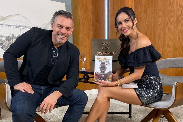 Luigi Baricelli de camisa social preta, sorrindo, sentado num banco branco ao lado de Daniela Albuquerque durante gravação do Sensacional, da RedeTV!