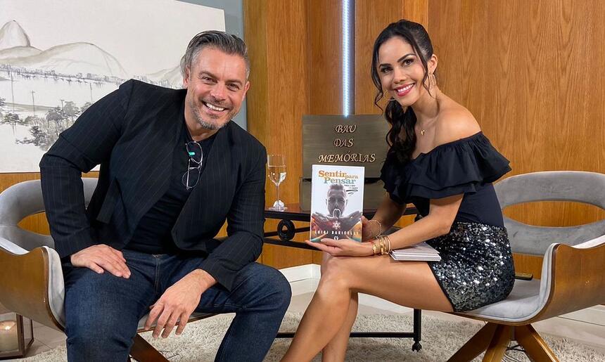 Luigi Baricelli de camisa social preta, sorrindo, sentado num banco branco ao lado de Daniela Albuquerque durante gravação do Sensacional, da RedeTV!