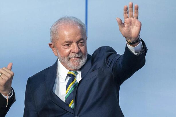 Lula com paletó preto, camisa social branca, gravata estampada em amarelo verde e azul, acenando com a mão esquerda durante debate