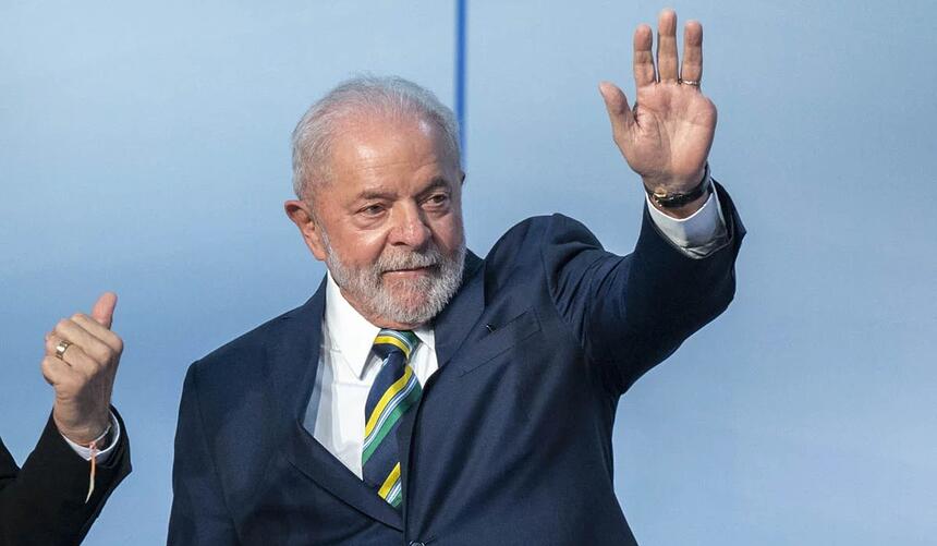 Lula com paletó preto, camisa social branca, gravata estampada em amarelo verde e azul, acenando com a mão esquerda durante debate