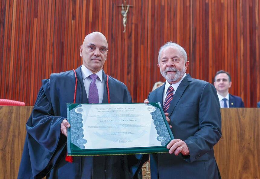 Lula e Alexandre de Moraes de blazer preto e camisa social branca, com gravata estampada vermelha, segurando diploma em cerimônia no TSE