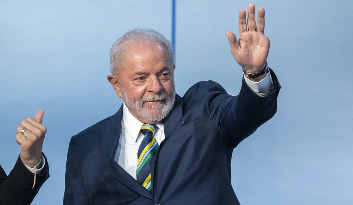 Lula com paletó preto, camisa social branca, gravata estampada em amarelo verde e azul, acenando com a mão esquerda durante debate