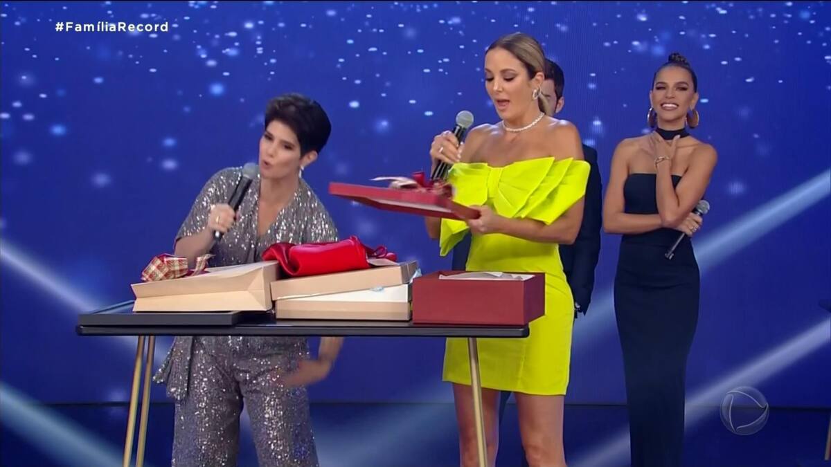 Mariana Godoy de macacão brilhante tira Ticiane de vestido amarelo no Família Record. Ao fundo de preto está Mariana Rios no palco do programa. 