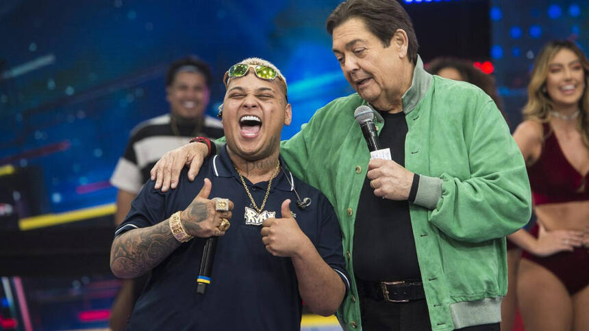 MC Ryan SP sorrindo, com a boca aberta, cabelo loiro, segurando o microfone com a mão direita; Faustão de jaqueta verde, camisa preta, segurando o microfone com a mão esquerda durante gravação de programa