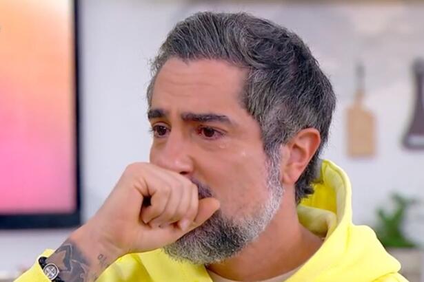 Marcos Mion de camisa de capuz amarelo, com a mão na boca, sentado num banco branco, chorando durante entrevista no Mais Você, da TV Globo