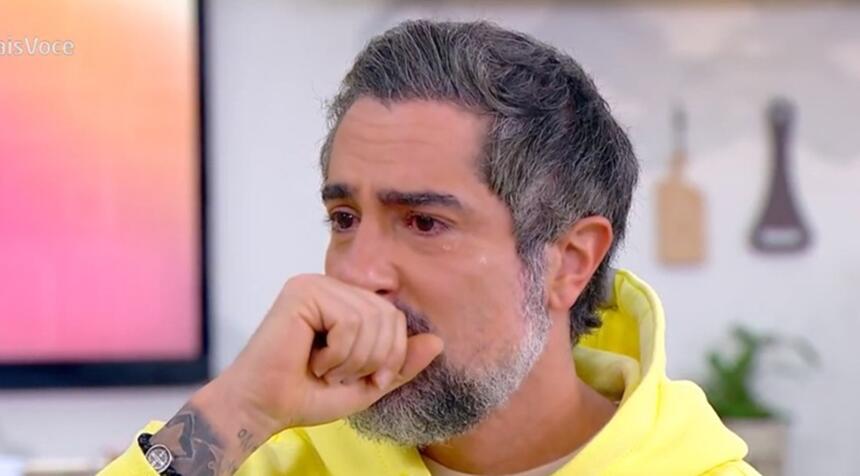 Marcos Mion de camisa de capuz amarelo, com a mão na boca, sentado num banco branco, chorando durante entrevista no Mais Você, da TV Globo