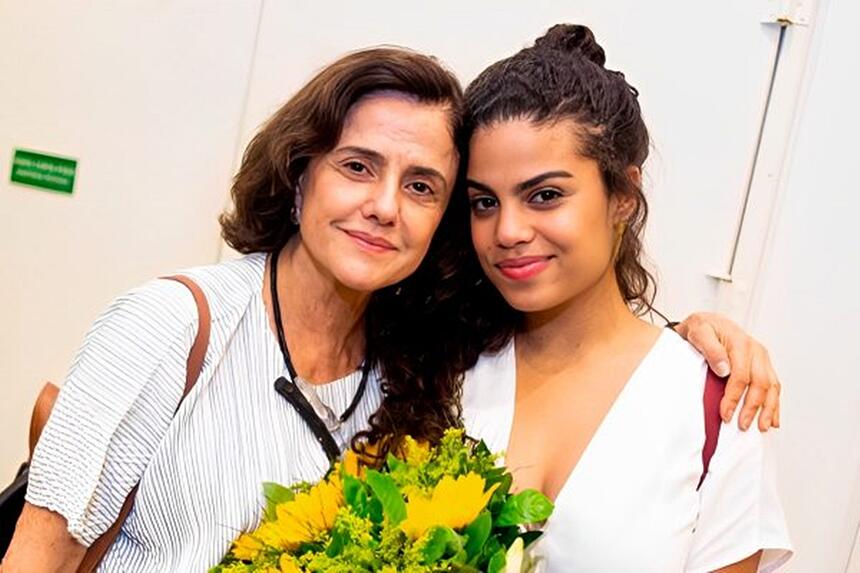 Marieta Severo de camisa branca, colar de madeira, segurando um buquê de flores amarelas ao lado de Clara Buarque, de cabelo preso, sorrindo tímidamente