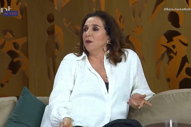 Marisa Orth, de roupa branca, sentada no sofá no estúdio do programa Saia Justa, no GNT