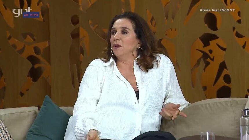 Marisa Orth, de roupa branca, sentada no sofá no estúdio do programa Saia Justa, no GNT