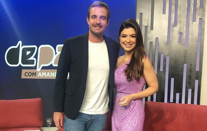 Max Fercondini de blazer azul marinho, camisa básica branca, sorrindo, ao lado de Amanda Françozo, que está de vestido lilás, sorrindo, durante gravação do "De Papo", da TV Aparecida