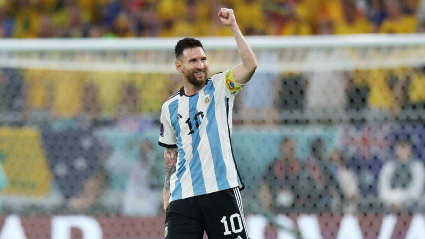 Messi com o sorrio aberto, em campo com a camisa da Argentina, em jogo da Copa do Mundo. Ele está com o braço esquerdo levantado