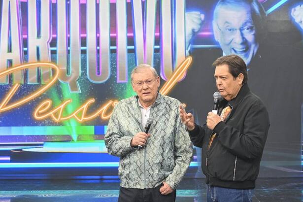 Milton Neves de camisa estampada em cinza e branco, calça preta, em pé, com óculo de grau, segurando o microfone com a mão direita durante gravação do 'Faustão na Band'