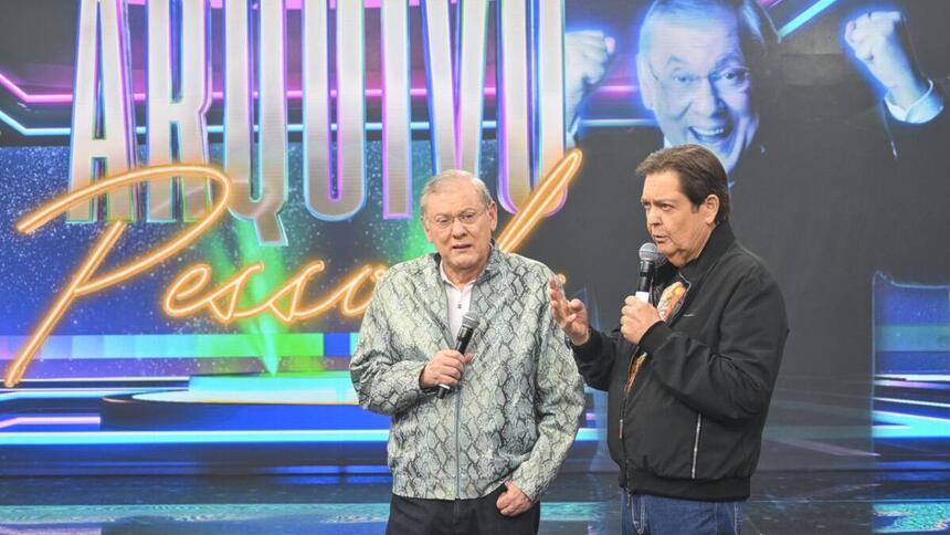 Milton Neves de camisa estampada em cinza e branco, calça preta, em pé, com óculo de grau, segurando o microfone com a mão direita durante gravação do 'Faustão na Band'