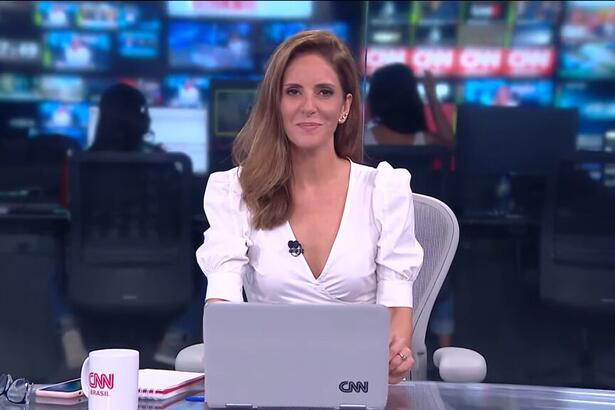 Monalisa Perrone, com uma blusa branca, sentada na bancada do Jornal da CNN, com um notebook a sua frente e a redação ao fundo