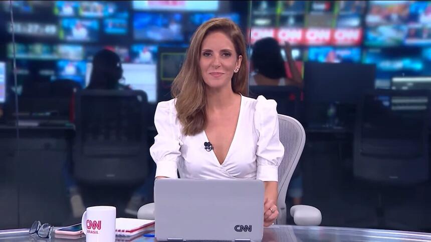 Monalisa Perrone, com uma blusa branca, sentada na bancada do Jornal da CNN, com um notebook a sua frente e a redação ao fundo