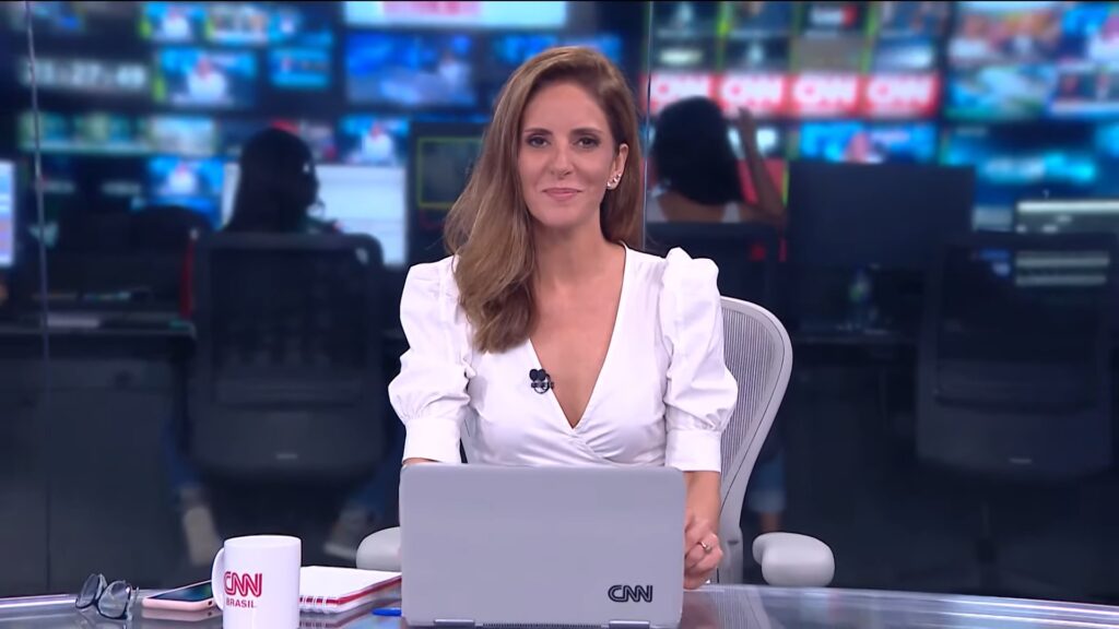 Monalisa Perrone, com uma blusa branca, sentada na bancada do Jornal da CNN, com um notebook a sua frente e a redação ao fundo