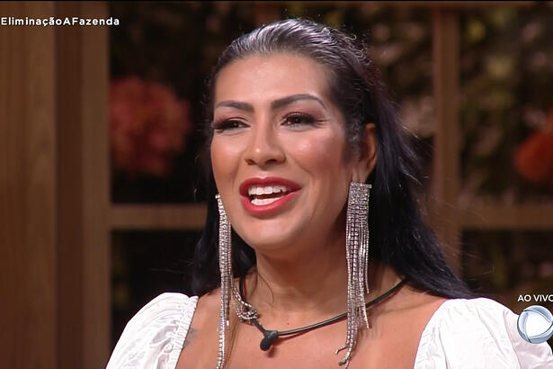 Moranguinho de vestido branco, com brinco de brilhantes, sorrindo, sentada na sede de eliminação de A Fazenda 14, da Record TV