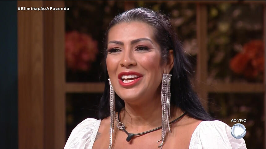 Moranguinho de vestido branco, com brinco de brilhantes, sorrindo, sentada na sede de eliminação de A Fazenda 14, da Record TV