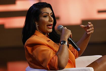 Moranguinho de blazer laranja, sentada num banco branco, segurando o microfone durante gravação do Hora do Faro, da Record TV