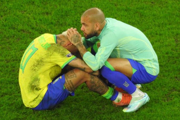 Neymar, sentado e de cabeça baixa, chorando em campo com a camisa da Seleção Brasileira, sendo consolado pro Daniel Alves