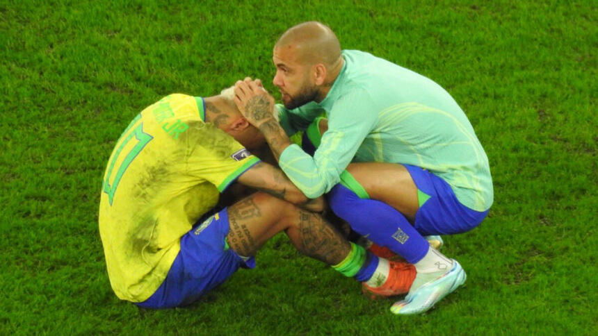 Neymar, sentado e de cabeça baixa, chorando em campo com a camisa da Seleção Brasileira, sendo consolado pro Daniel Alves