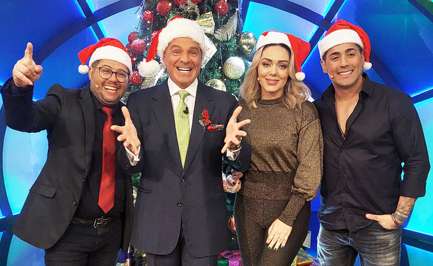 Tânia Mara de camisa e calça em tons de marrom, sorrindo, com gorro de papai noel ao lado de Marcello de Carvalho e a dupla Hugo e Tiago que estão de camisa e calça preta, sorrindo, também com gorro