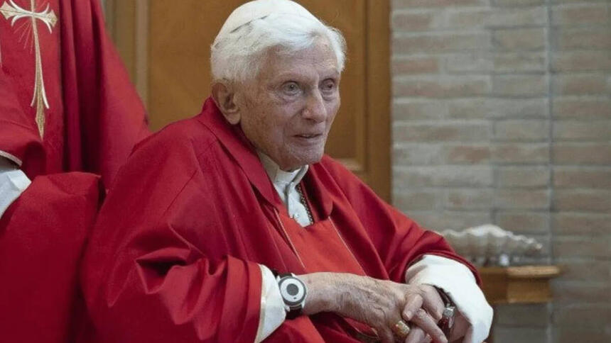 Papa Bento XVI, de batina, sentado em um evento da Igreja Católica