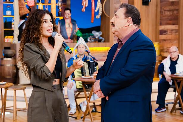 Paula Fernandes de macaquito verde musco, segurando o microfone com a mão direita, usando cabelo ondulado, solto e top preto, em pé, ao lado de Ratinho; Ratinho de blazer azul, camisa social lilás, com os braços juntos durante gravação do Boteco, do SBT