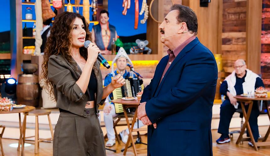 Paula Fernandes de macaquito verde musco, segurando o microfone com a mão direita, usando cabelo ondulado, solto e top preto, em pé, ao lado de Ratinho; Ratinho de blazer azul, camisa social lilás, com os braços juntos durante gravação do Boteco, do SBT