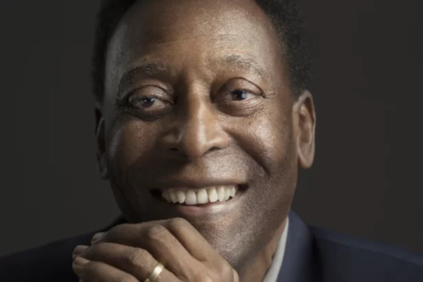 Pelé sorrindo, de blazer preto e camisa social branca, com as mãos encostadas no rosto durante ensaio fotográfico