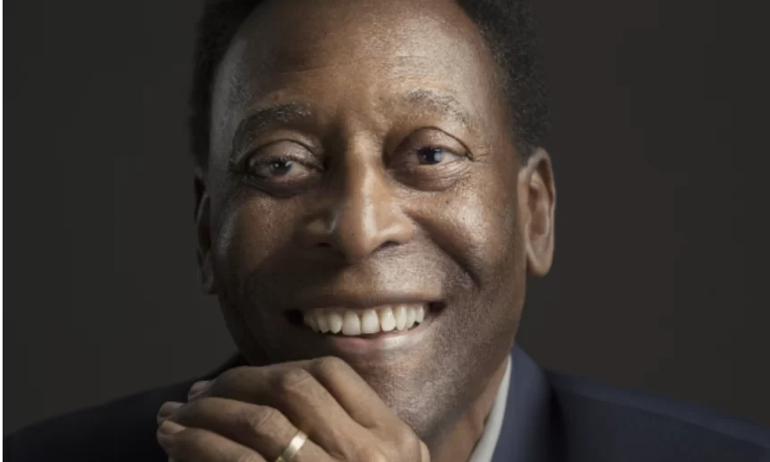 Pelé sorrindo, de blazer preto e camisa social branca, com as mãos encostadas no rosto durante ensaio fotográfico