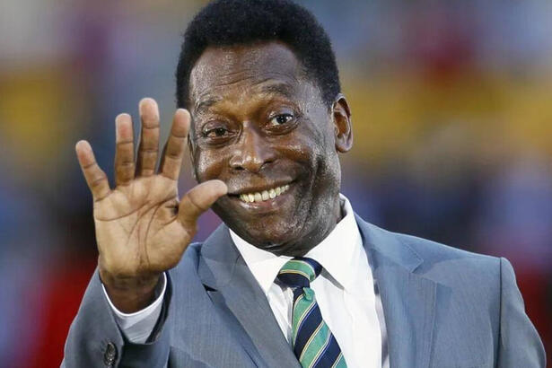 Pelé, de terno e gravata, com um sorriso no rosto, tendo o braço levantando e saudando