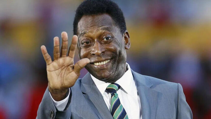 Pelé, de terno e gravata, com um sorriso no rosto, tendo o braço levantando e saudando