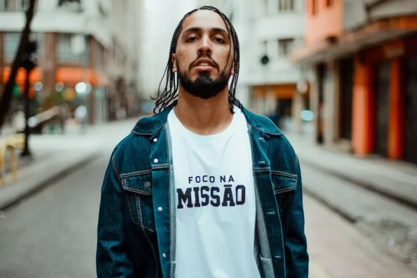 Rashid de jaqueta jeans, camisa branca, de cabelo trnaçado, andando pela rua fotografando para divulgação de álbum