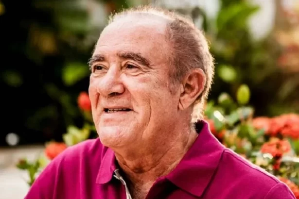 Renato Aragão de camisa polo vinho, de perfil, sorrindo, num jardim