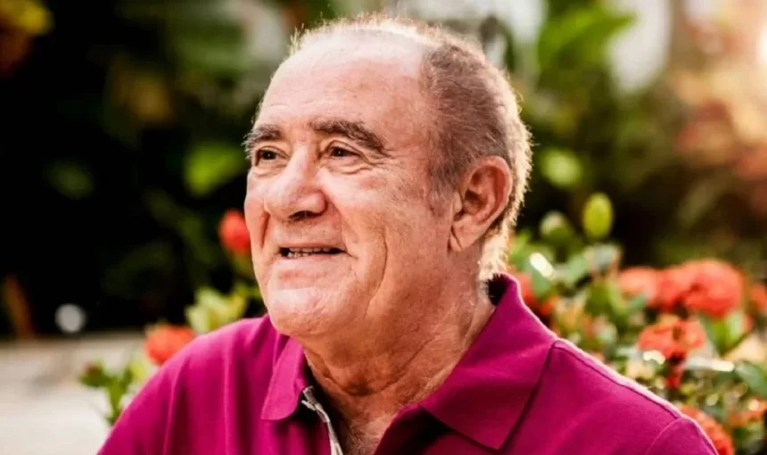 Renato Aragão de camisa polo vinho, de perfil, sorrindo, num jardim