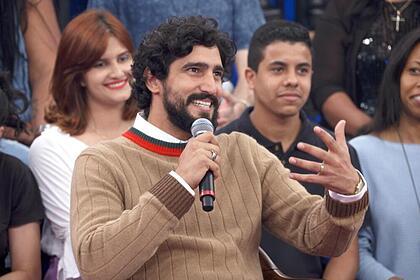 Renato Góes de cardigã marrom, camisa vermelha e branca, sorrindo, segurando um microfone e gesticulando com a mão durante gravação do Altas Horas, da TV Globo