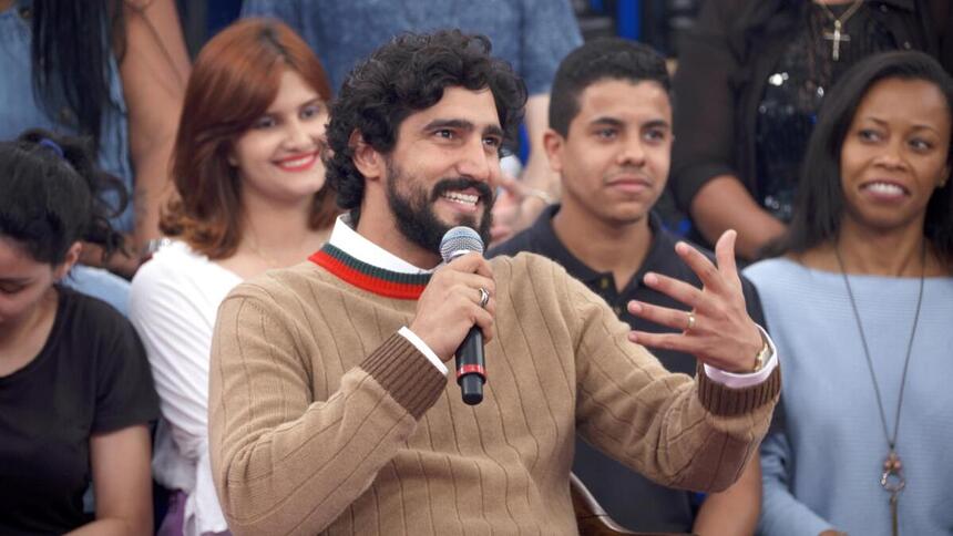 Renato Góes de cardigã marrom, camisa vermelha e branca, sorrindo, segurando um microfone e gesticulando com a mão durante gravação do Altas Horas, da TV Globo