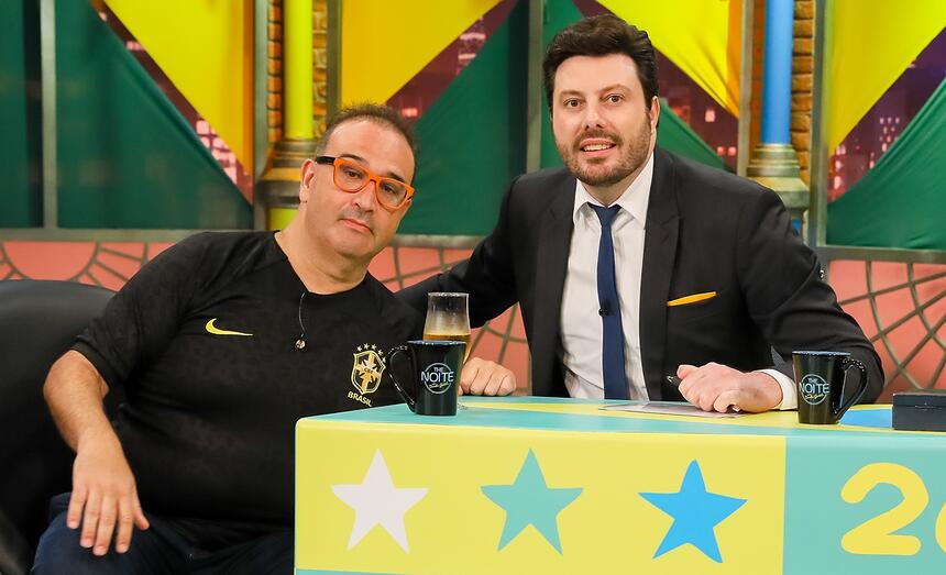 Renato Tortorelli de camisa da Seleção Brasileira preta, de óculos de grau, sentado num sofá branco ao lado de Danilo Gentili, durante gravação do The Noite, do SBT