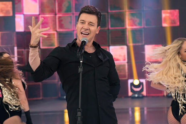 Rodrigo Faro de camisa e calça brancos com a mão pra frente homenageando Ricky Martin durante gravação do seu programa na Record TV