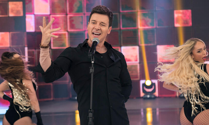 Rodrigo Faro de camisa e calça brancos com a mão pra frente homenageando Ricky Martin durante gravação do seu programa na Record TV