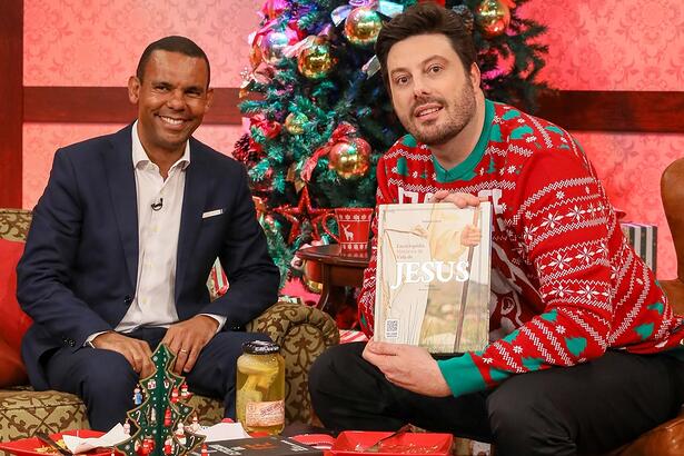 Rodrigo Silva de blazer azul, camisa social branca, calça jeans escura, sentado num sofá ao lado de uma árvore de Natal ao lado de Danilo Gentili, que está sorrindo, sentado, segurando um livro durante gravação do The Noite, do SBT